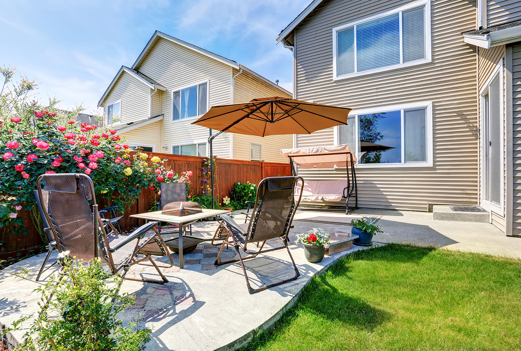 A sunny backyard patio with lounge chairs, a table, and a large tan umbrella next to a blooming pink rose bush, a green lawn, and a house with beige siding.