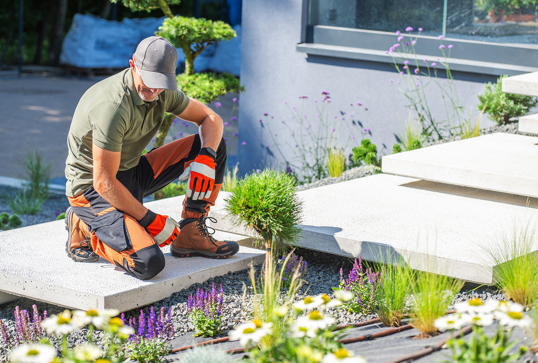 A professional gardener in orange and black work trousers and a cap kneels on a modern concrete slab, tending to a newly landscaped garden with purple flowers, ornamental grasses, and a drip irrigation system.