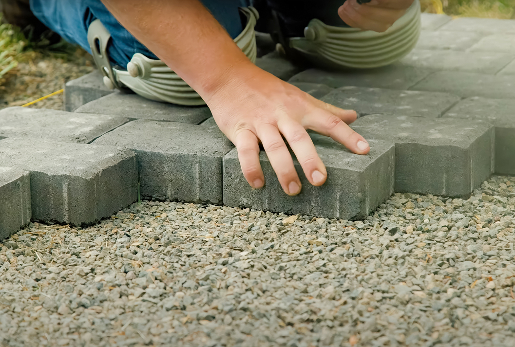 A close-up of a person wearing knee pads laying grey interlocking pavers on a bed of gravel for a patio or walkway installation.
