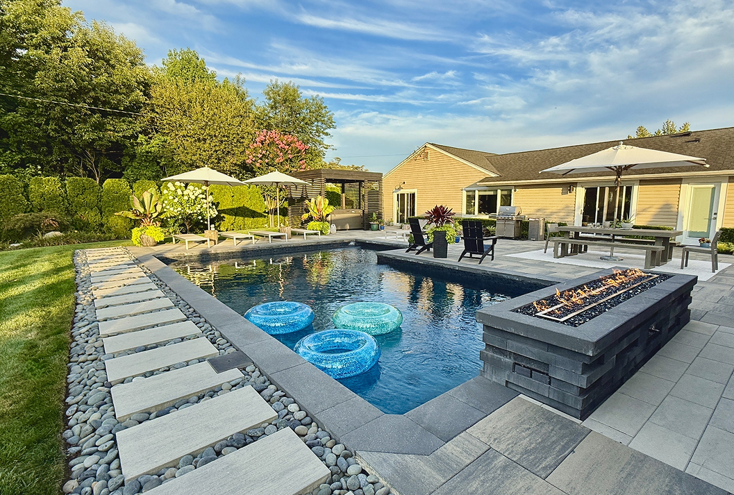 A luxurious backyard with a modern rectangular swimming pool, a lit stone fire pit, and a large paver patio featuring outdoor dining and lounge areas with umbrellas, all set against a lush green landscape and a tan house under a blue sky.
