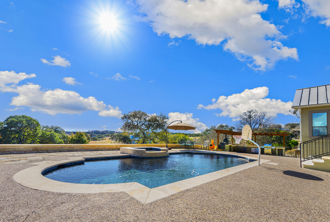 A sunlit backyard patio with a dark-bottom swimming pool, attached hot tub, and a poolside basketball hoop, overlooking the rolling hills on a bright, sunny day.