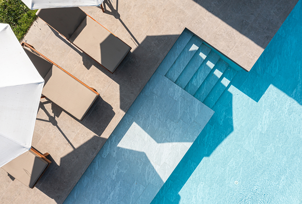A bird's-eye view of a modern luxury poolside, featuring a turquoise swimming pool with stone steps, and a tiled patio with lounge chairs and white umbrellas casting strong geometric shadows in the sun.