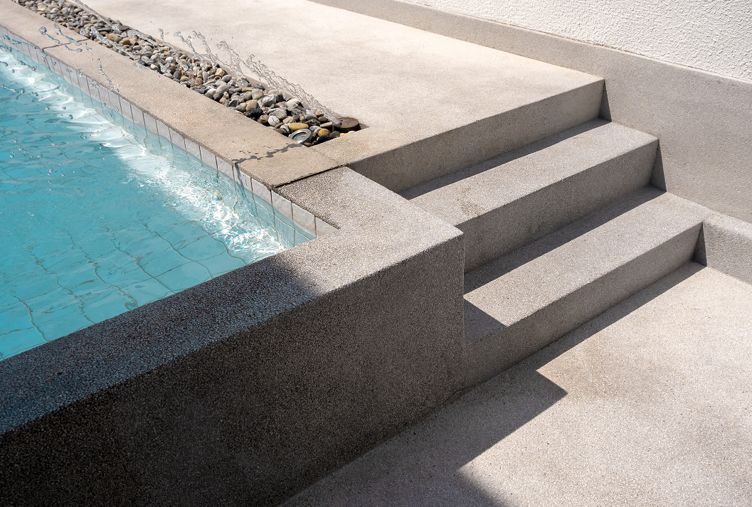 A modern, architectural swimming pool corner with minimalist concrete steps and a textured exposed aggregate finish, featuring a water feature with splashing water over river pebbles alongside the blue-tiled pool.