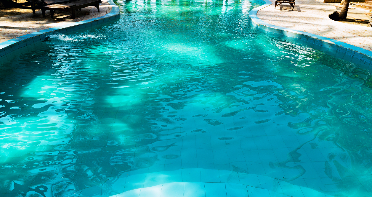 Bright turquoise outdoor swimming pool with sunlit ripples and tree shadows, surrounded by a tiled deck with wooden lounge chairs.