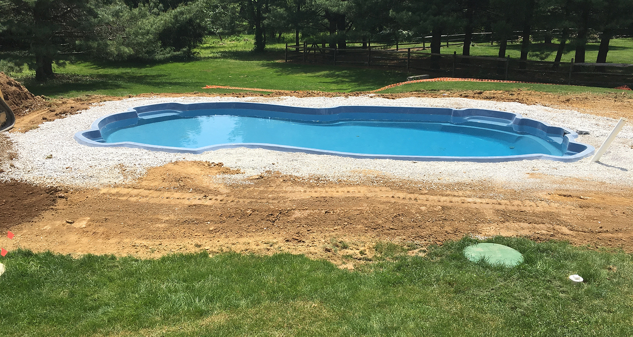 Installation of a freeform light blue fiberglass in-ground swimming pool in a backyard surrounded by white gravel and construction soil.