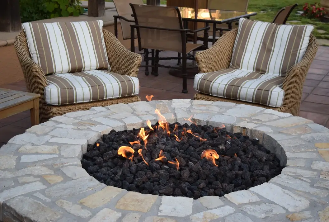 Outdoor stone fire pit with burning flames and comfortable wicker patio chairs.
