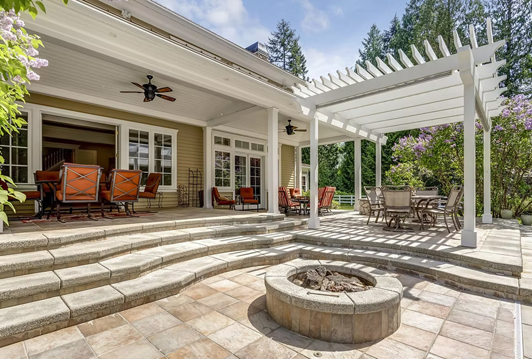 A luxurious multi-level stone patio with a round, built-in fire pit on the lower level and wide, curved steps leading up to a covered porch. The outdoor living space includes multiple seating areas and a white pergola over a dining set, all surrounded by lush green trees and flowering shrubs.