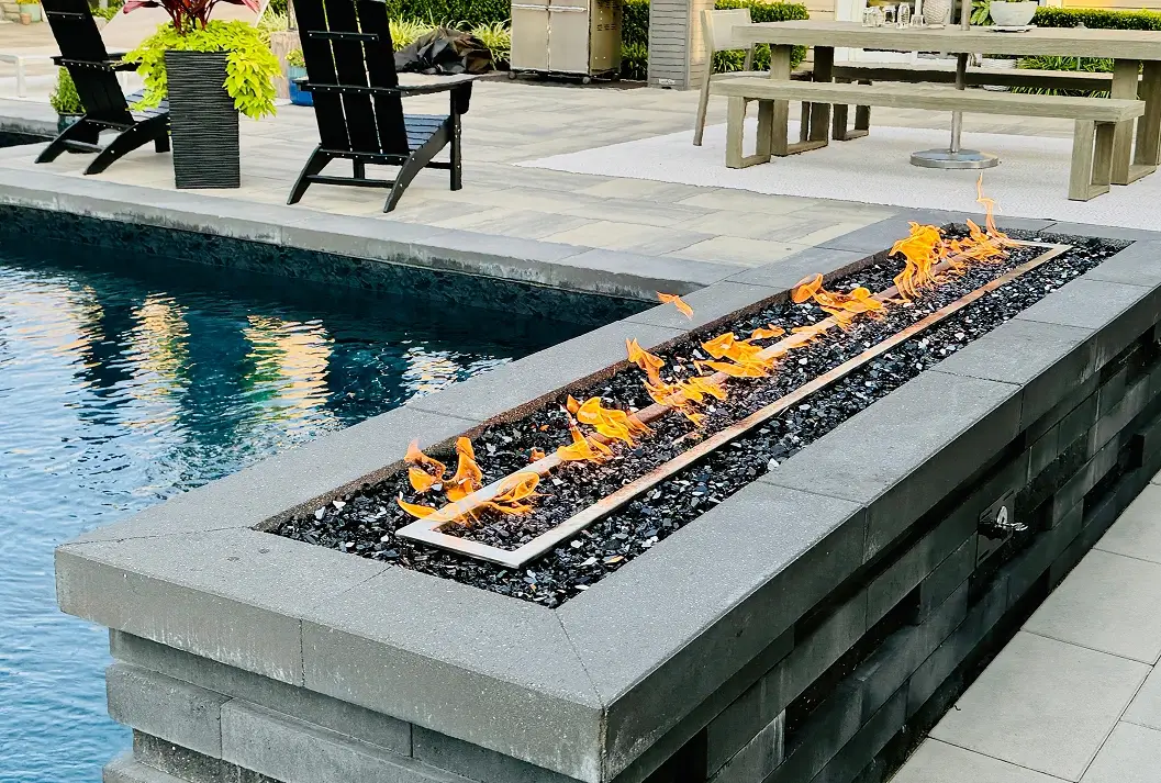 Modern linear gas fire pit with flames next to a backyard swimming pool.