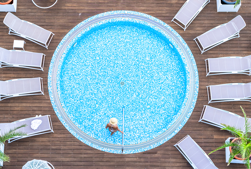 Top-down aerial view of a woman in a straw hat and bikini relaxing in a round, blue mosaic-tiled swimming pool on a wooden deck surrounded by empty sun loungers.