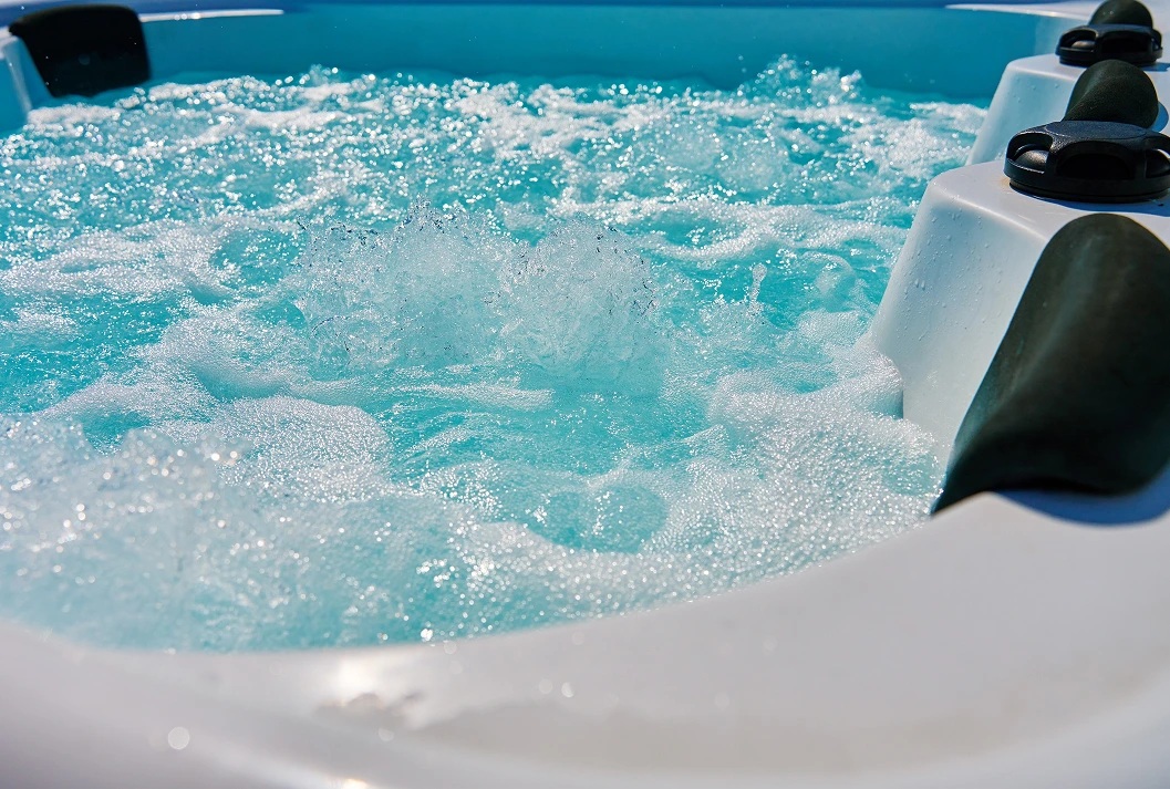 A close-up view of a white hot tub with clear, turquoise water bubbling vigorously from the jets, creating white foam under bright sunlight.