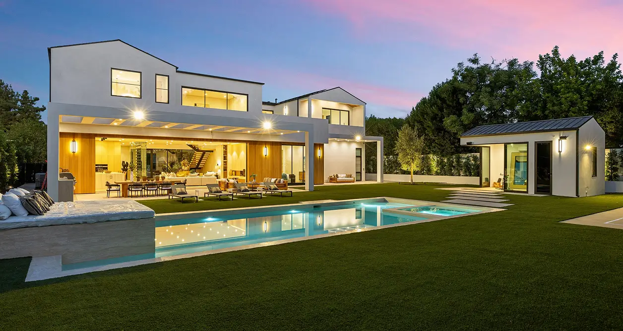 Modern luxury house at dusk with illuminated swimming pool, hot tub, and lawn.