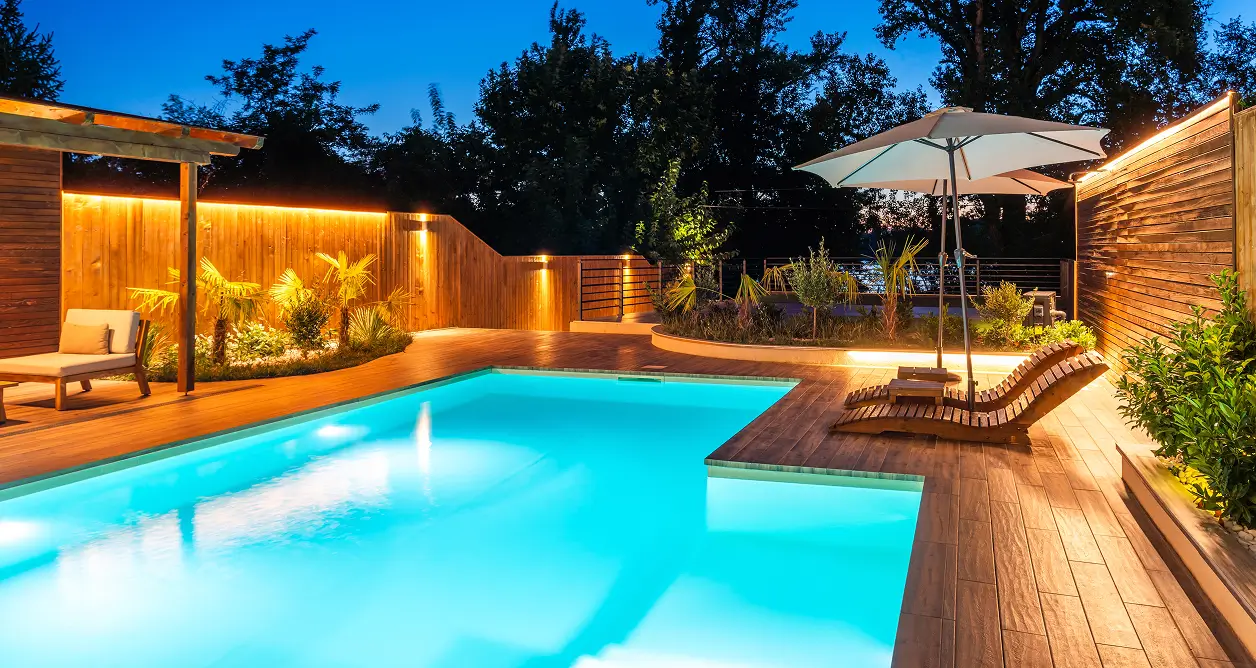 Backyard pool with warm LED deck lighting, wooden patio, and loungers at night.