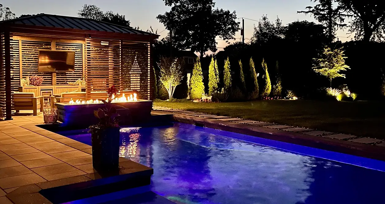 Illuminated backyard pool with fire pit, gazebo, and landscape lighting at night.