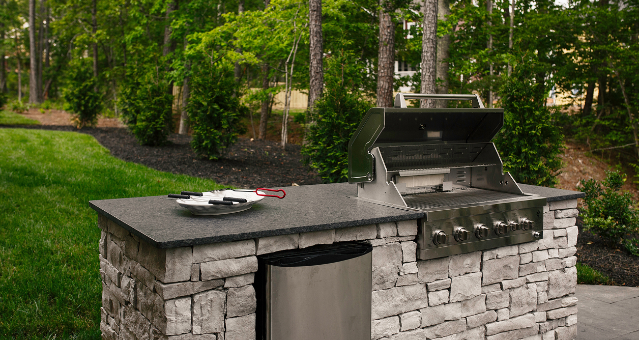 A custom-built outdoor kitchen island made of gray stacked stone with a dark granite countertop, featuring a built-in stainless steel gas grill with an open lid and a mini-refrigerator, set in a landscaped backyard with a green lawn and a wooded area in the background.