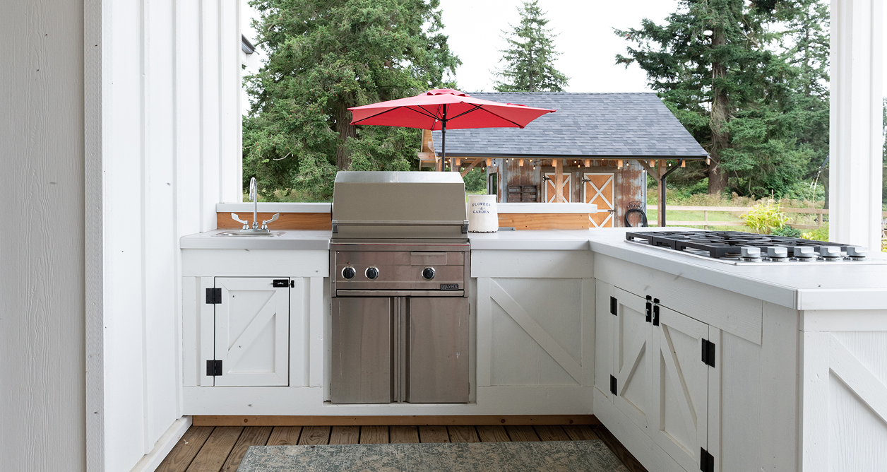 A modern farmhouse-style outdoor kitchen on a covered porch, featuring white cabinets with black barn door hardware, a built-in stainless steel BBQ grill, a gas cooktop, and a small sink. A red umbrella is open behind the grill, with a view of a rustic barn and trees.