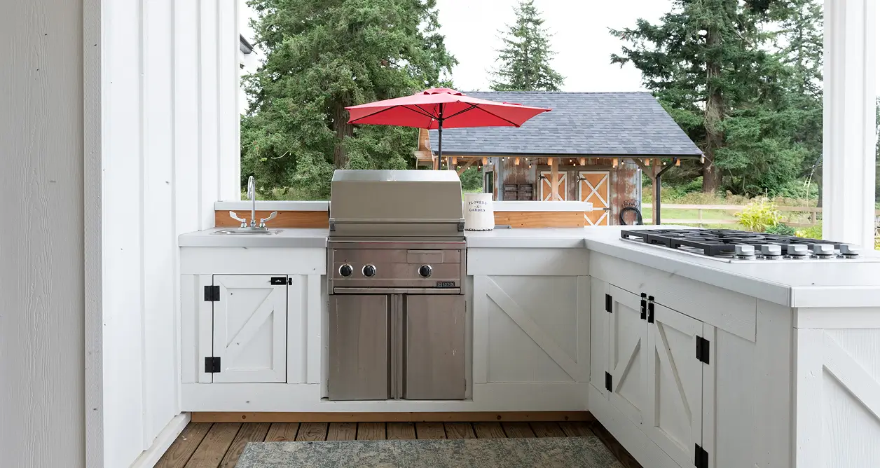 White farmhouse-style outdoor kitchen with stainless steel grill, stovetop, and sink.