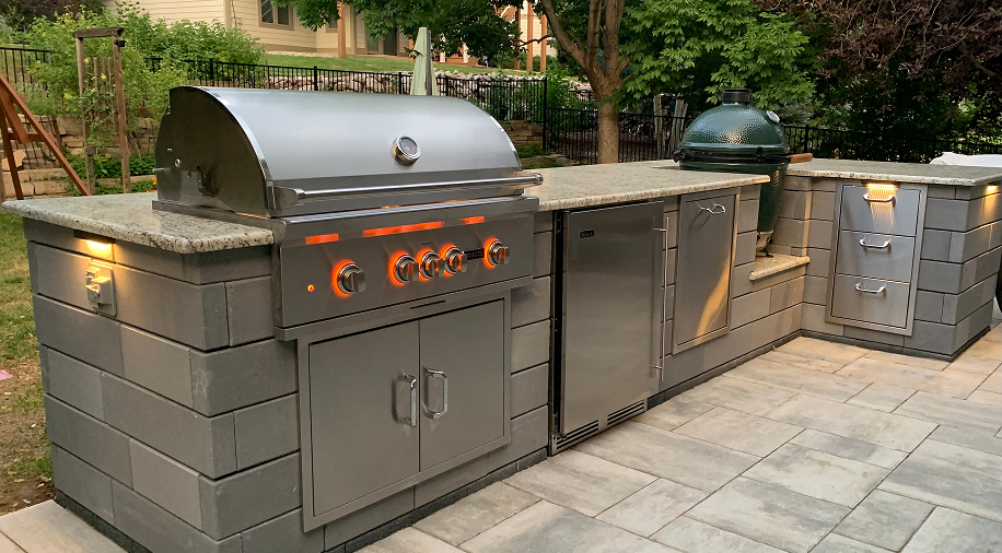 Custom outdoor kitchen featuring a stainless steel gas grill with illuminated knobs, a green kamado ceramic grill, built-in stainless steel refrigerator, and granite countertops on a grey stone base and paved patio.