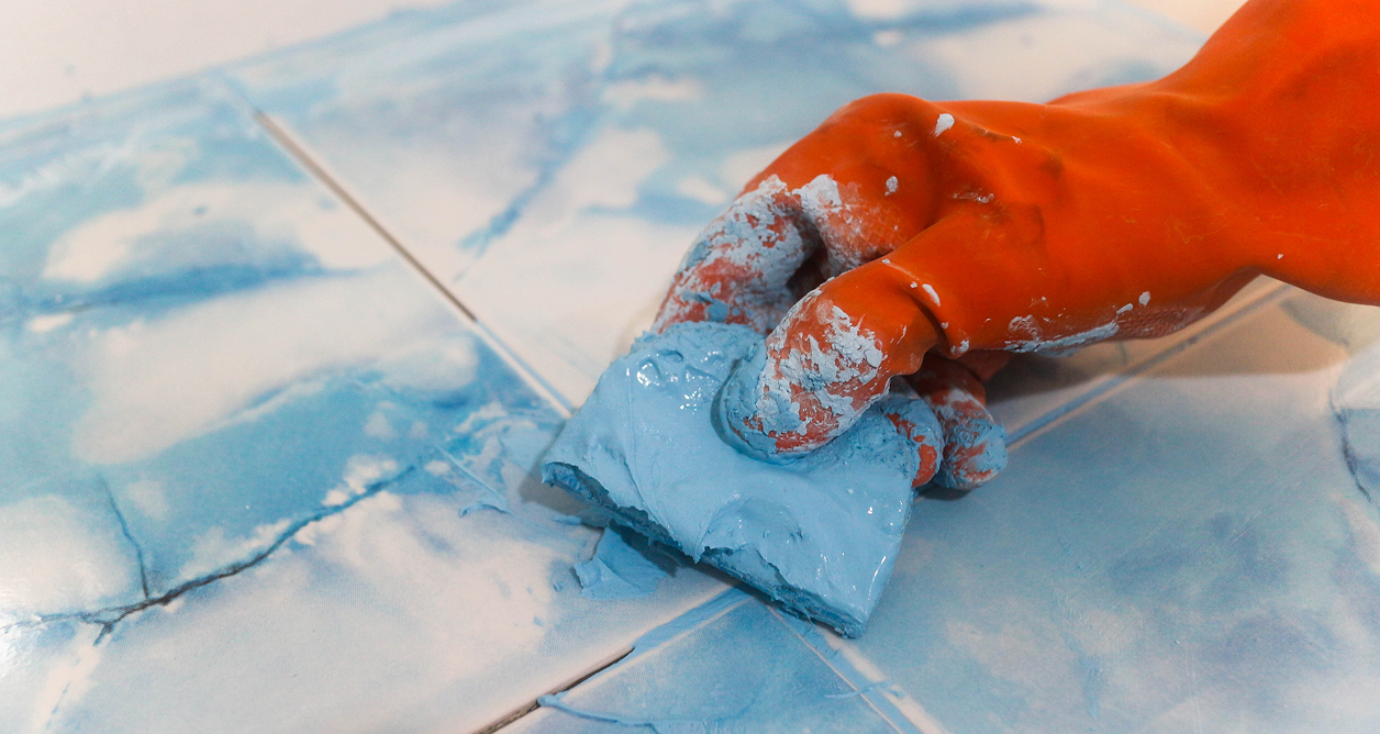 Hand in orange glove applying blue grout to marble pattern tiles.