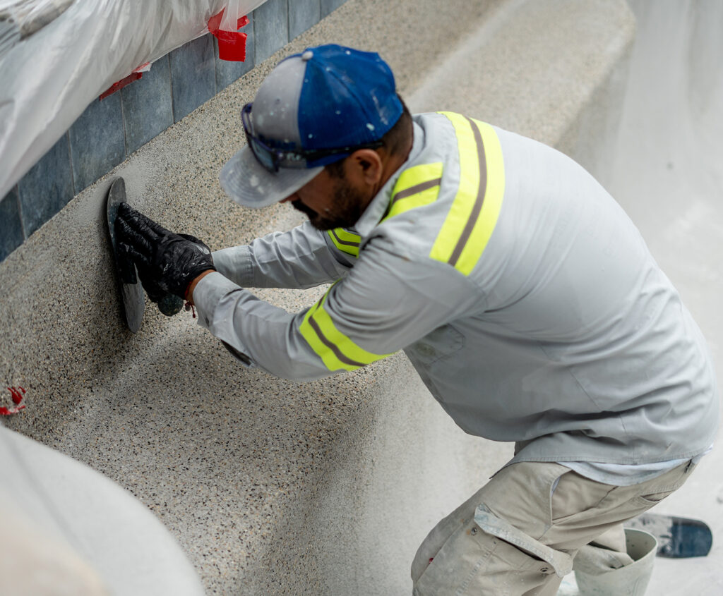 Professional worker applying pebble finish to a swimming pool wall with a trowel during a pool renovation project.