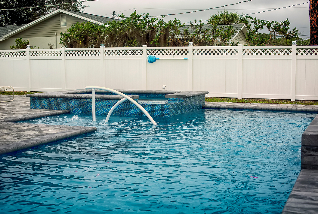 A backyard swimming pool with an integrated spa featuring blue mosaic tiles and a white handrail, set against a white vinyl privacy fence with a lattice top. The pool is surrounded by a grey paver deck.