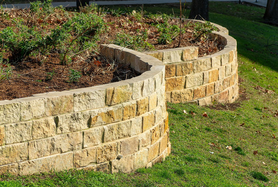 A curved, tiered retaining wall made of rough-hewn tan and yellow stone blocks creates a raised garden bed on a sloping green lawn.