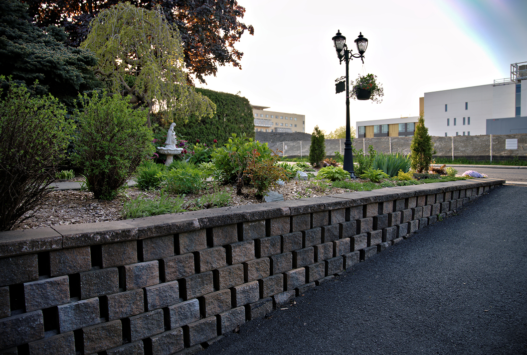 A low-angle shot of a stacked concrete block retaining wall bordering a landscaped garden with a vintage lamppost, a weeping tree, and a small statue, with modern buildings and a faint rainbow in the background.