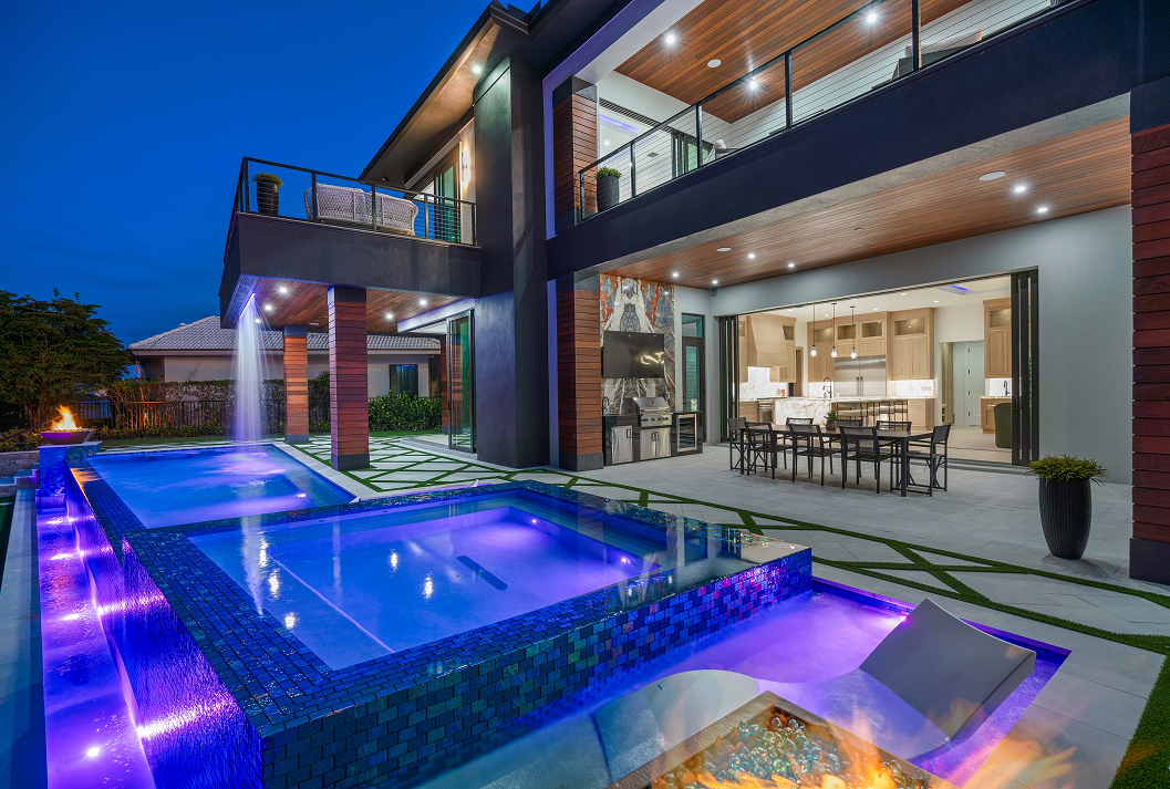 A luxurious two-story modern home at dusk, featuring a stunning backyard patio and a multi-level swimming pool with vibrant blue LED lighting. The pool includes an integrated spa, a waterfall cascading from an upper balcony, and a fire pit. The covered outdoor living space has a dining area and a built-in kitchen, which opens into the house through large sliding glass doors, creating a seamless indoor-outdoor experience.