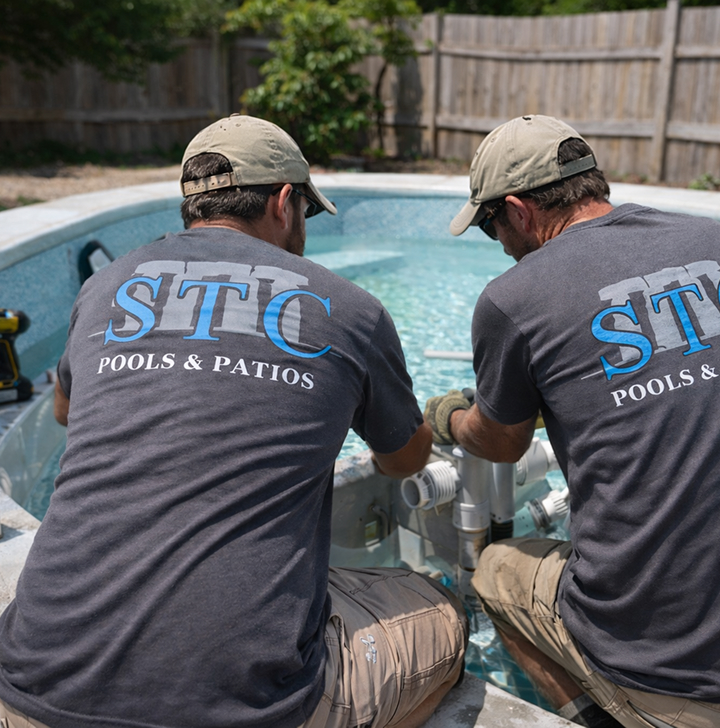 Two STC Pools & Patios technicians in matching grey logo t-shirts kneel beside a residential swimming pool, working together to install new plumbing and equipment.