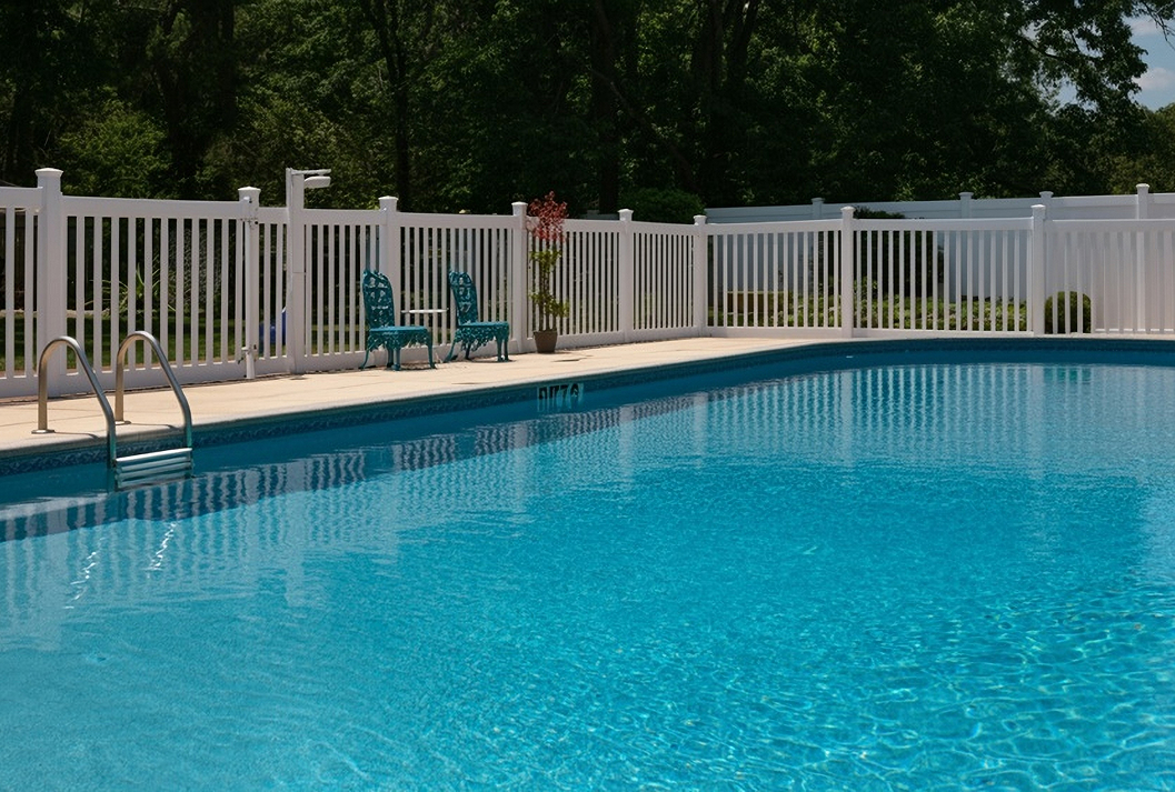 A white vinyl picket fence surrounding a sparkling blue in-ground swimming pool with a pool ladder and two teal patio chairs on the concrete deck.