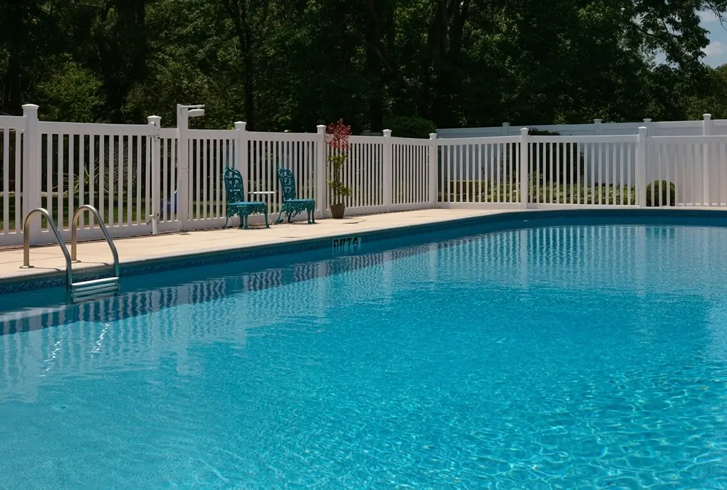 A white vinyl picket fence surrounding a sparkling blue in-ground swimming pool with a pool ladder and two teal patio chairs on the concrete deck.