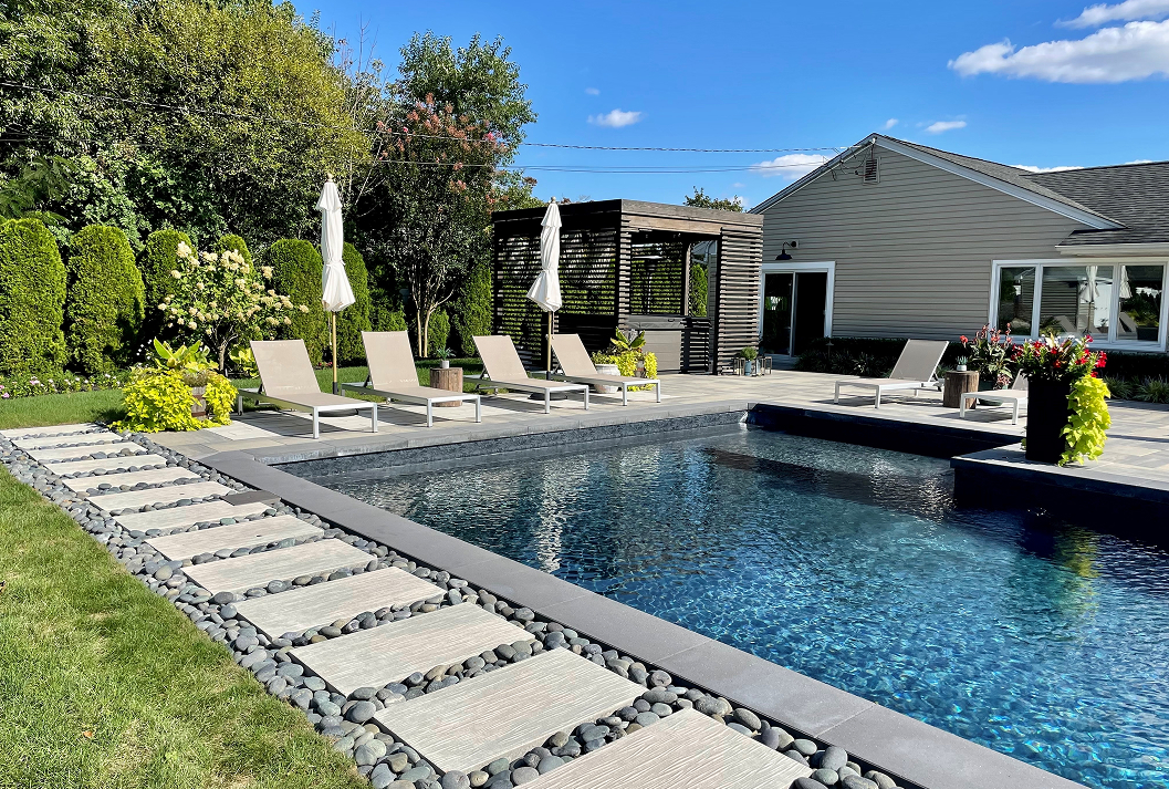 A modern, landscaped backyard on a sunny day featuring a rectangular in-ground swimming pool with dark blue water. A stone path with river rocks borders the pool and a green lawn. The patio includes beige lounge chairs, white umbrellas, and a contemporary dark wood pergola next to a grey house.