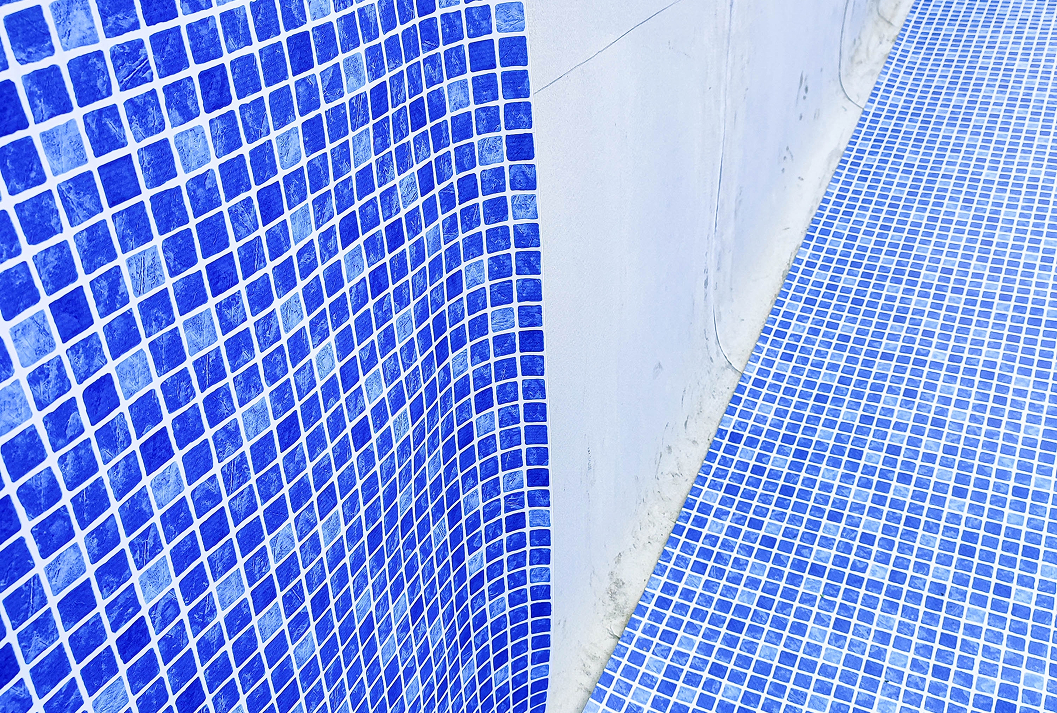 A close-up, angled shot of the inside corner of an empty swimming pool, with the floor and one curved wall covered in small, blue and light blue mosaic tiles with white grout.