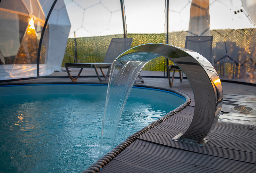 A contemporary chrome waterfall feature cascades into a round swimming pool on a dark wood deck, with a transparent geodesic dome and lounge chairs visible in the warm sunset glow.