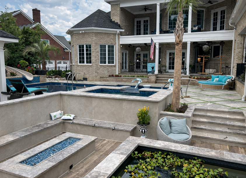 Luxurious backyard with a multi-level pool and spa, water slide, built-in fire pit, lily pad pond, modern lounge seating, and a large brick house with balconies.