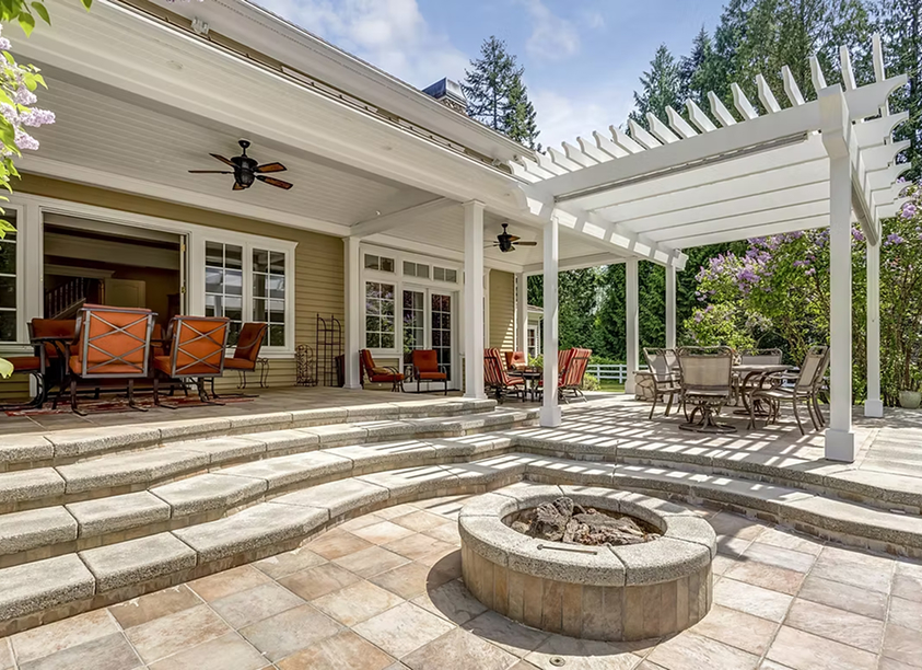 Back patio of a tan house with a white pergola, outdoor dining area, and a stone fire pit.