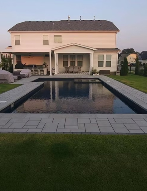 Modern backyard with a rectangular swimming pool and a light-colored patio, adjacent to a two-story house with a covered patio area and outdoor kitchen.