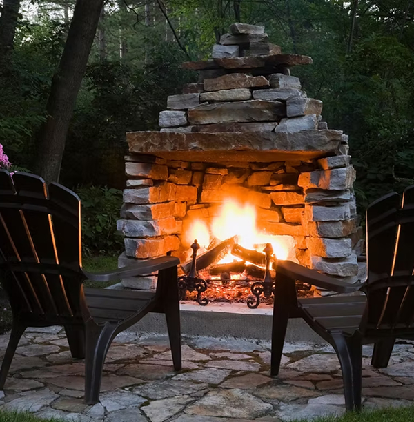 Stone outdoor fireplace with a crackling fire and two Adirondack chairs facing it on a flagstone patio.