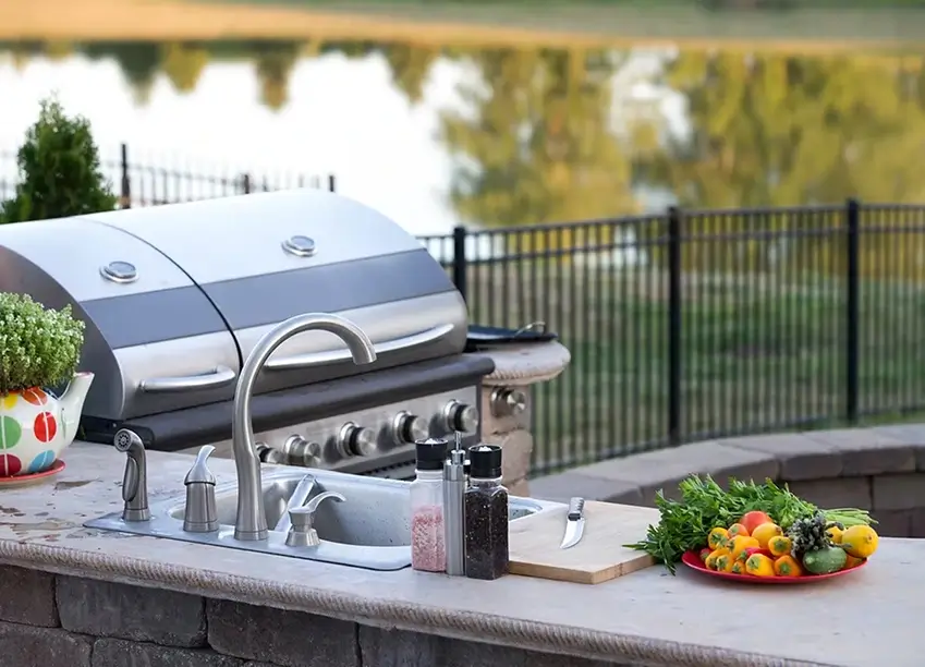 Outdoor kitchen island with stainless steel grill, sink, faucet, salt and pepper shakers, cutting board with knife, and a plate of colorful vegetables, with a fence and reflective water in the background.