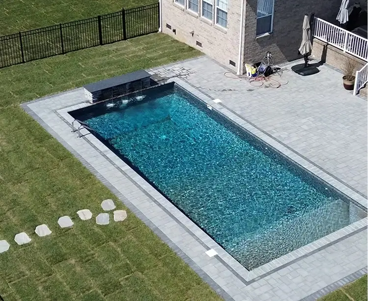 Rectangular swimming pool with dark blue water and built-in waterfall feature, surrounded by a grey paver patio and green lawn with stepping stones.