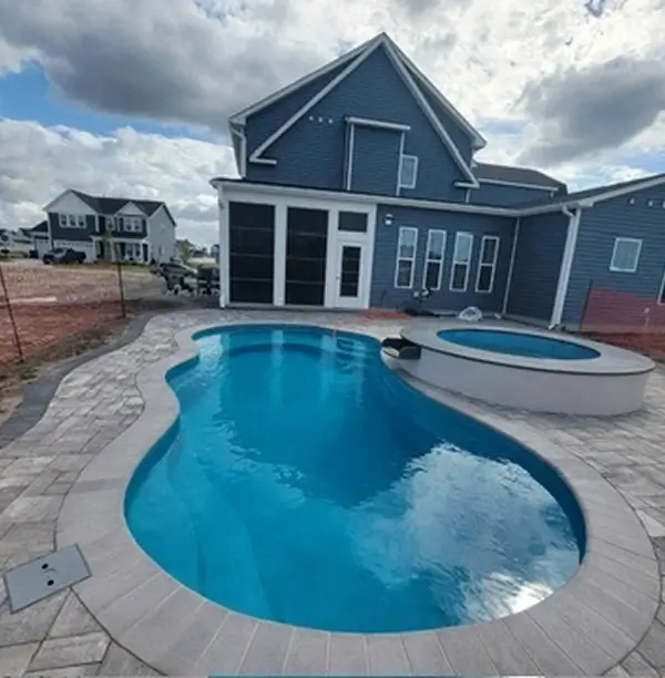 Blue swimming pool and spa next to a blue house with white trim and windows, surrounded by a stone patio and walkway.