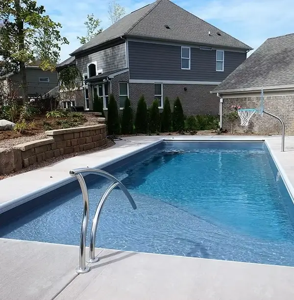 Rectangular swimming pool with blue water, surrounded by a concrete patio and a backyard with a house, landscaping, and a basketball hoop.